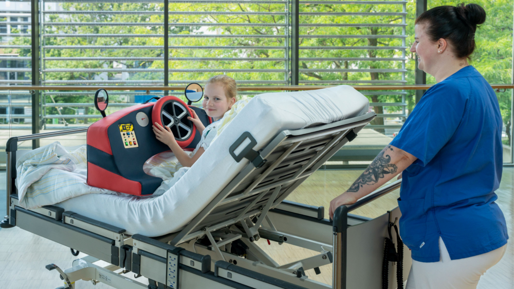 Schwester Katrin schiebt Greta in ihrem Patientenbett über den Krankenhausflur. Das mobile Cockpit lenkt Greta dabei ab und beschäftigt sie. ©UKD/Michael Kretzschmar