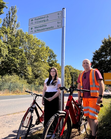 Auszubildende der Stadtverwaltung Coswig, Monique Liebetrau, und Steffen Wendler vom Baubetriebshof stehen an einem der neu angebrachten Radwegweiser an der Dresdner Straße. Foto: Stadtverwaltung Coswig